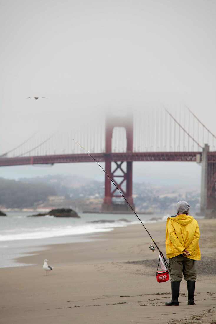 Man on beach fishing in front of Golden Gate Bridge covered with fog, San Francisco, USA