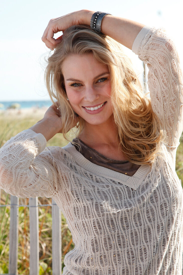 Portrait of pretty blonde woman wearing jumper with v neck standing on beach, smiling