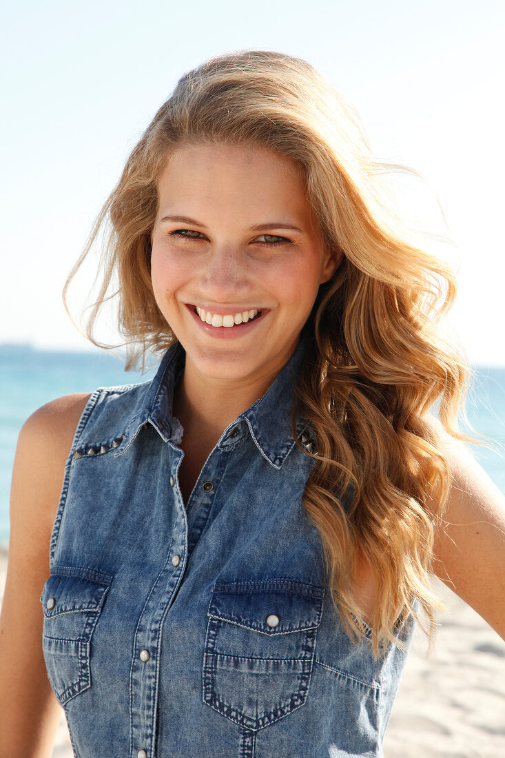 blonde Frau in blauem Jeanshemd am Strand, lacht im Kamera
