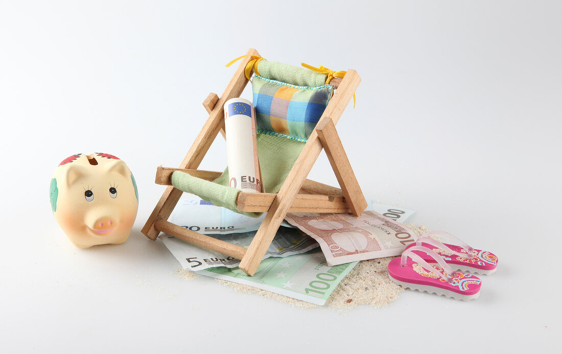Rolled bill on deck chair, flip-flops, sand and piggy bank on white background