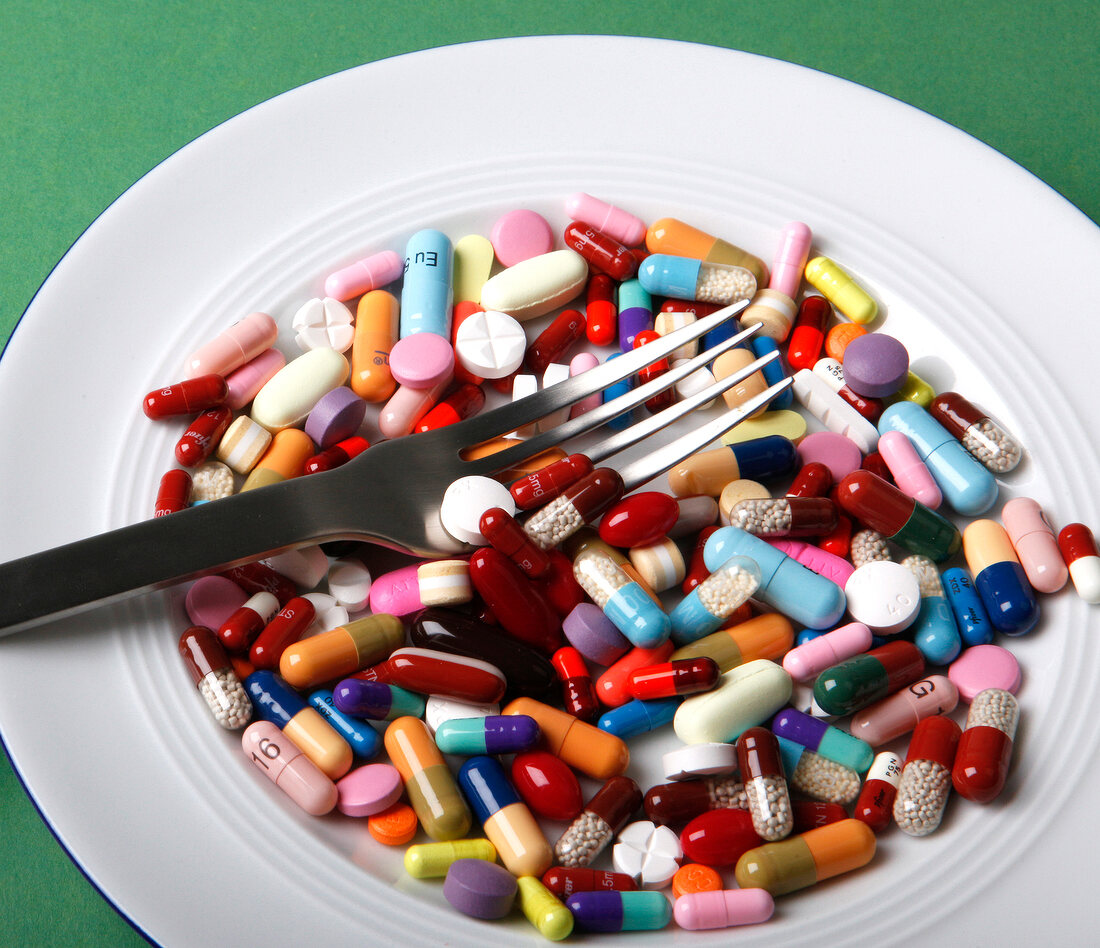 Different pills, tablets and capsules on plate with fork against green background