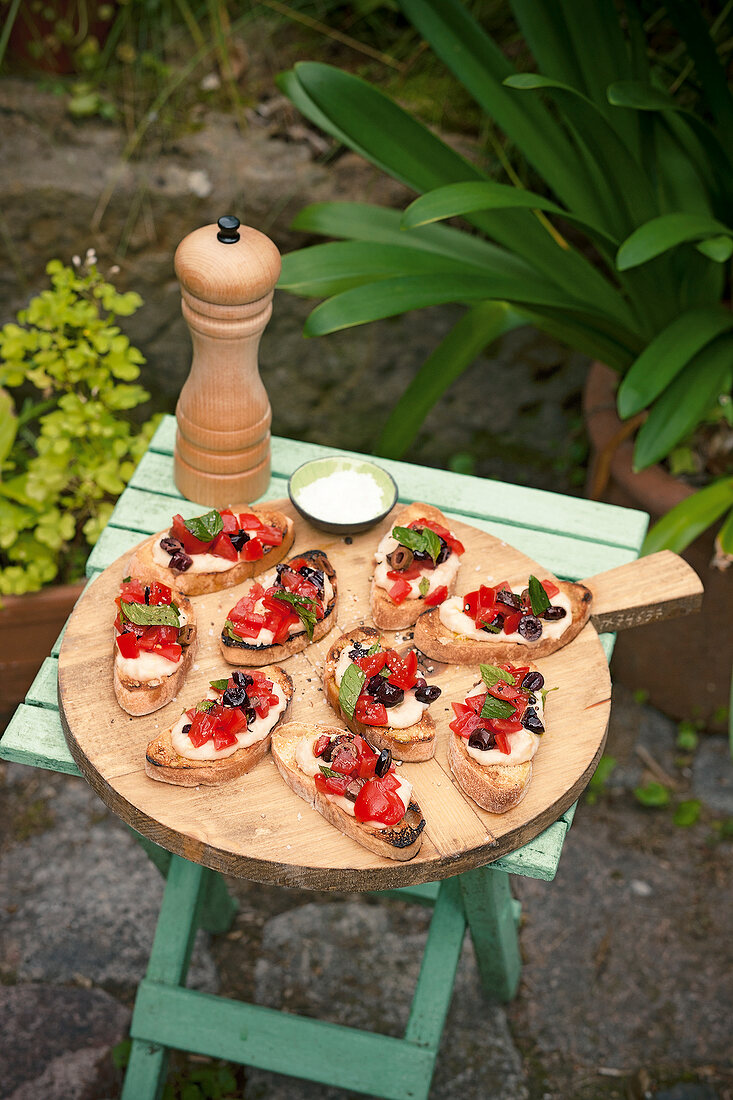 Bruschetta with tomatoes and olives on wooden tray, black pepper mill on stool