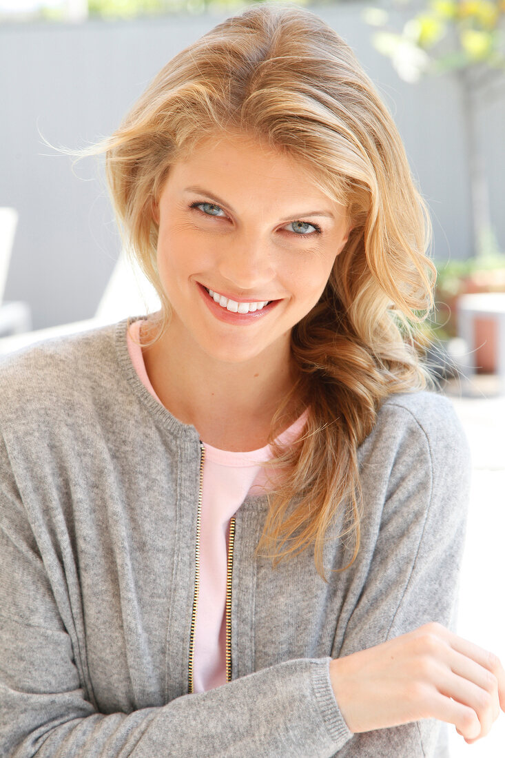 Portrait of blonde woman wearing pink top and gray jacket standing at poolside, smiling