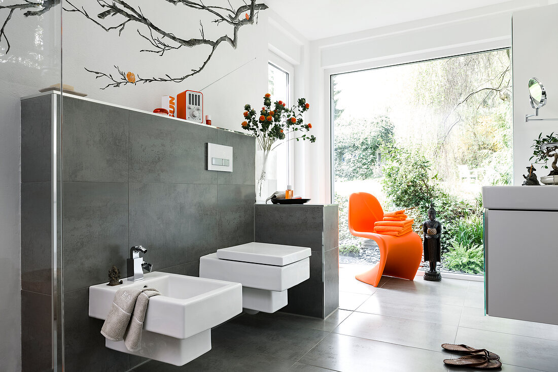 View of bathroom with white toilet seat, bidet and large window with orange chair