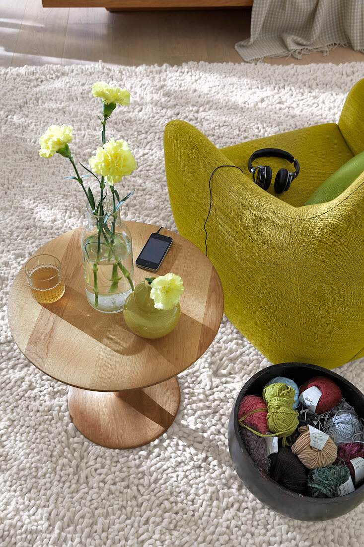 Basket with balls of wool and wooden table with glass flower vase besides green armchair