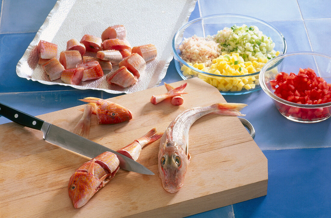 Sliced fish with knife on wooden cutting board and other ingredients in bowl on table