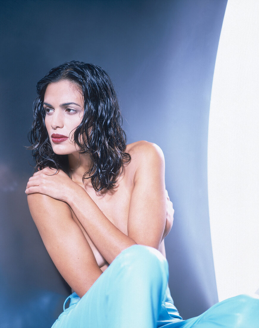 Semi-nude woman with dark wavy hair sitting with arms on her bare chest, looking away