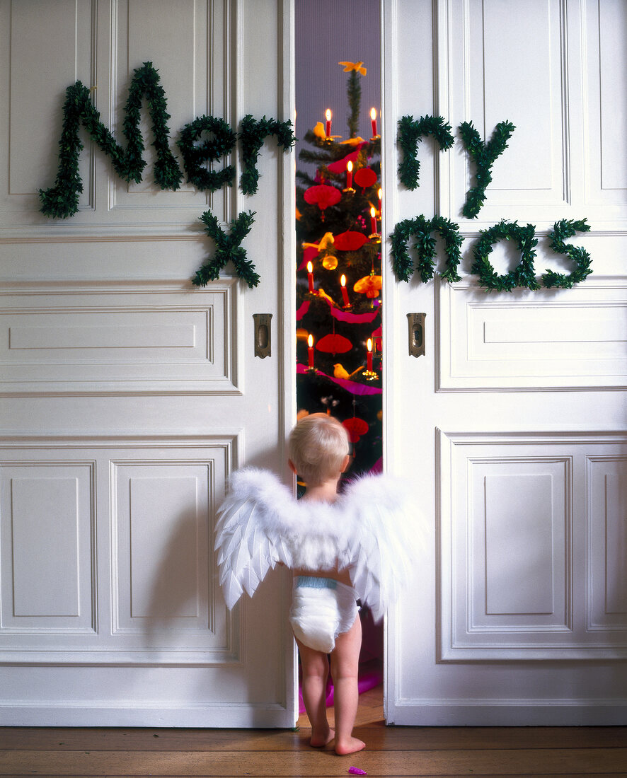 Baby als Engel verkleidet steht vor Merry X-Mas Tür (Windel, Flügel)