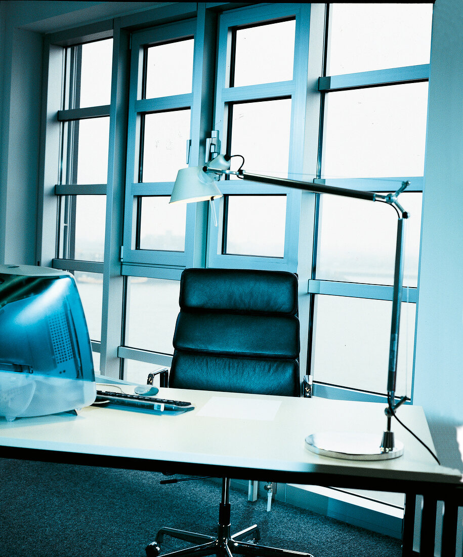 Desk with computer, black leather chair and lamp against window