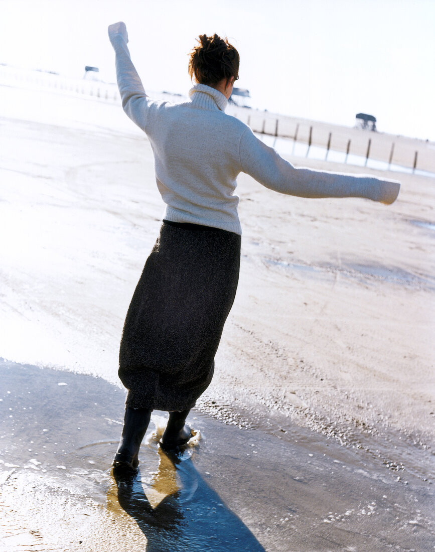 Rear view of woman in sweater walking on beach with arms out stretched