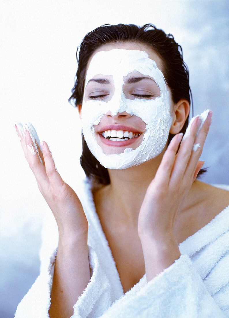 Happy woman in bathrobe applying white exfoliating cream on face, smiling and eyes closed