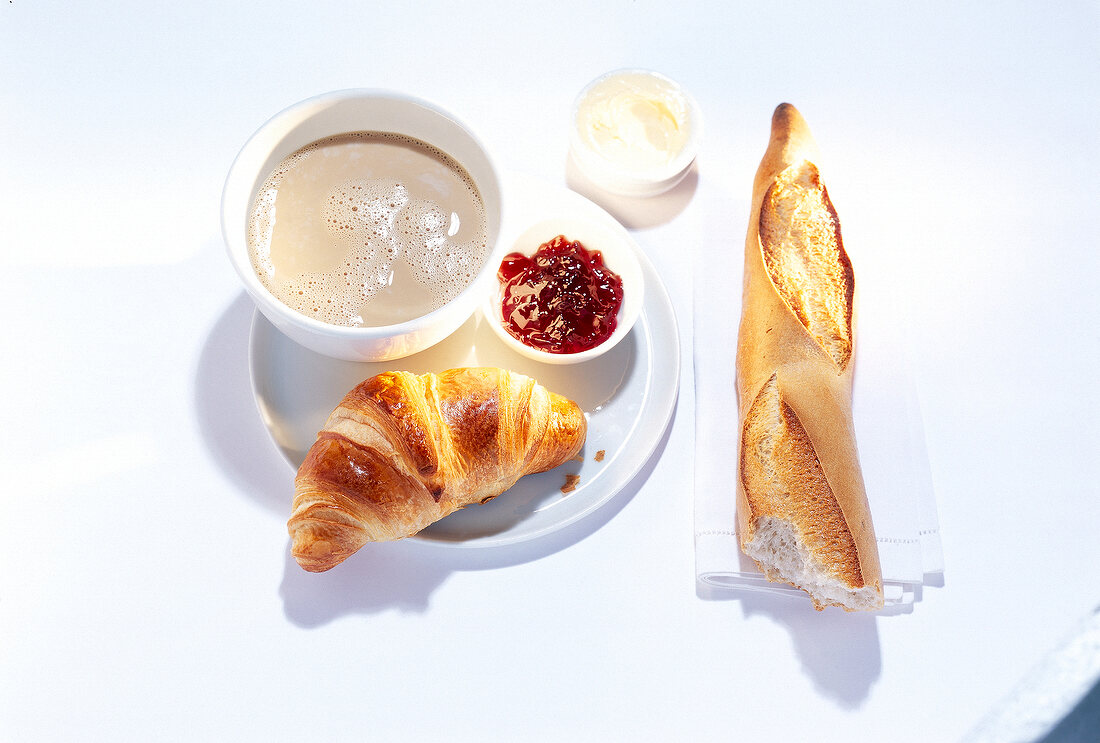 Breakfast table set with baguette, jam, croissant and coffee with milk on plate