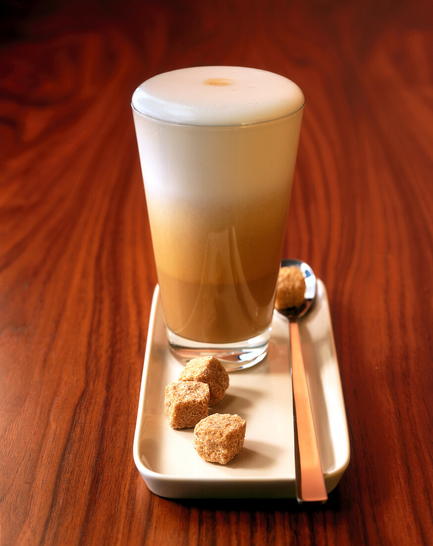 Latte macchiato in glass on white plate with sugar cubes on wooden surface