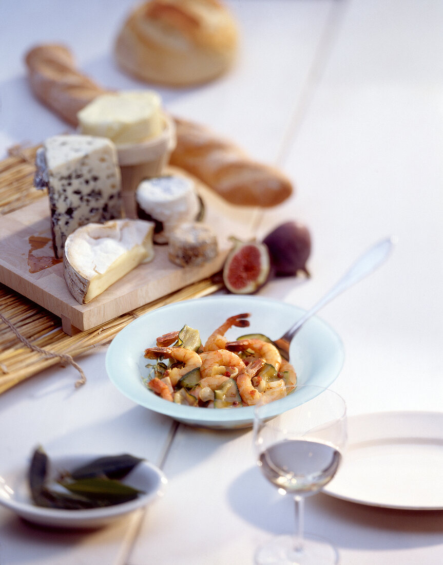 Plate of shrimp dish, cheese on wooden board with baguette and white wine on table