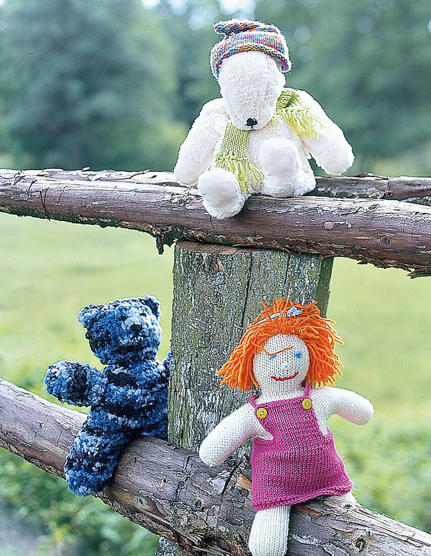Drei Strickfiguren: zwei Teddybären, eine Puppe auf einem Holzzaun