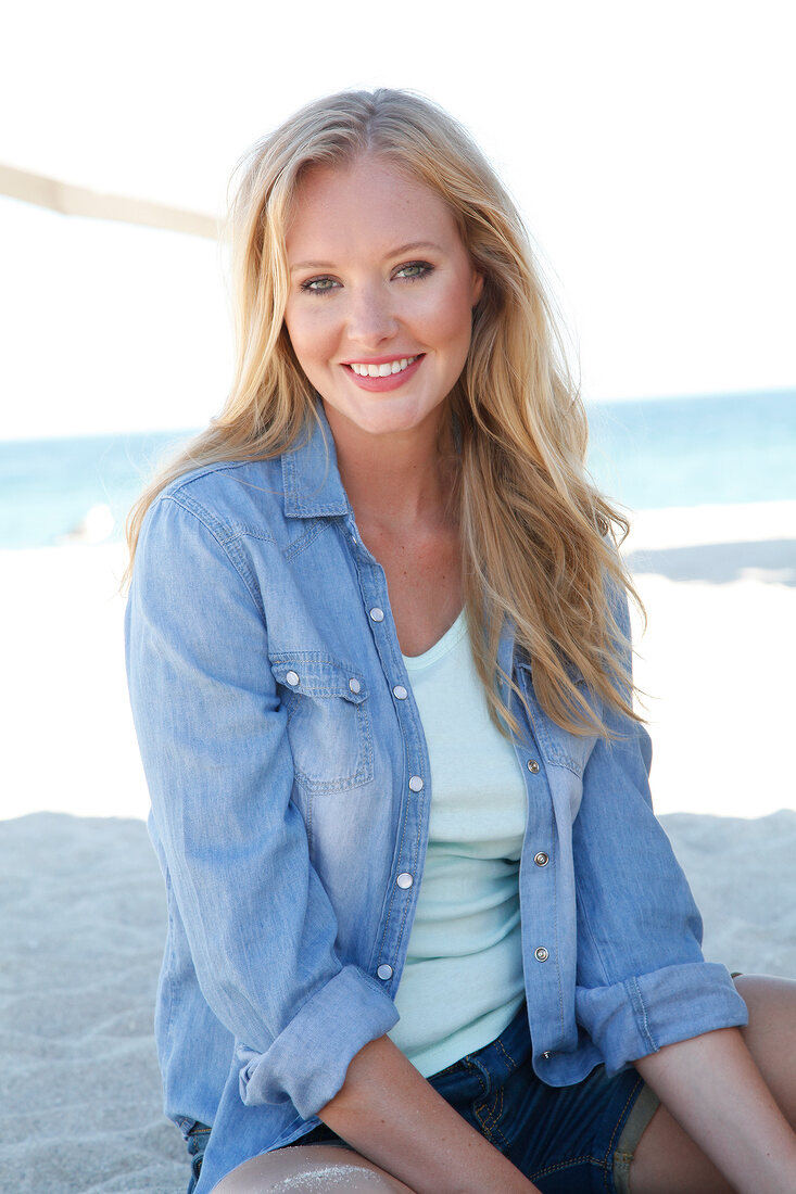 Blonde woman in a denim shirt smiles at the camera on the beach