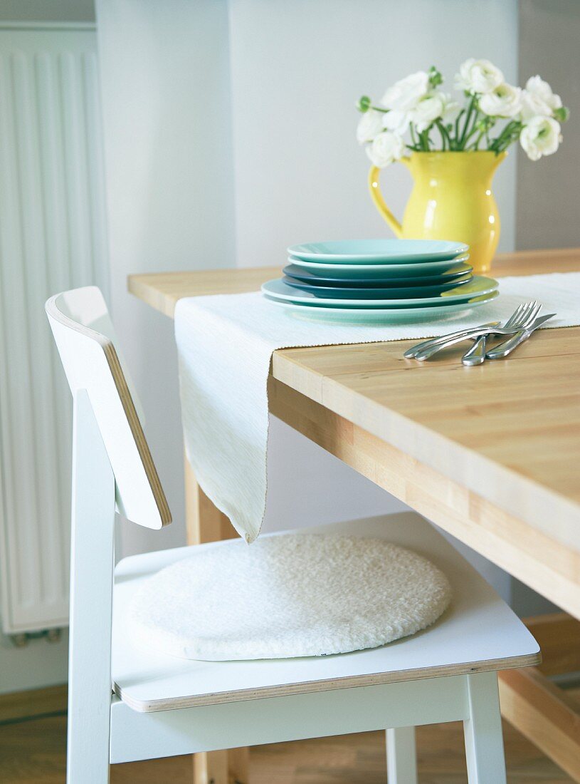 A stack of plates and a bunch of flowers in a yellow jug on a light table runner solid wooden table with a simple wooden chair in the foreground