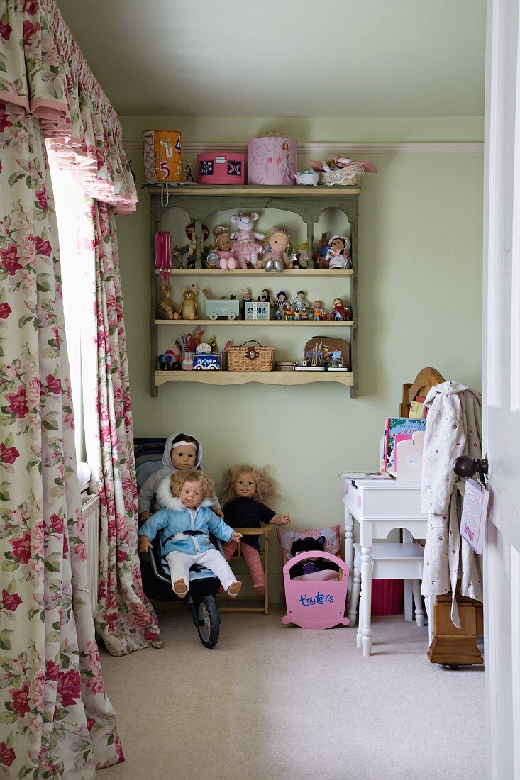 View through open door of nostalgic child's bedroom with dolls in buggy, toys on wall-mounted shelves and rose-patterned curtains with pelmet