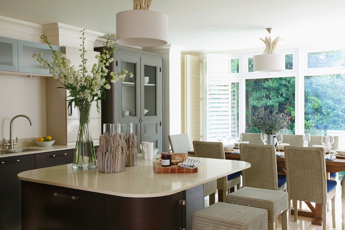 Open-plan kitchen in elegant, maritime, country-house style with rounded square island and wicker chairs at set table in background