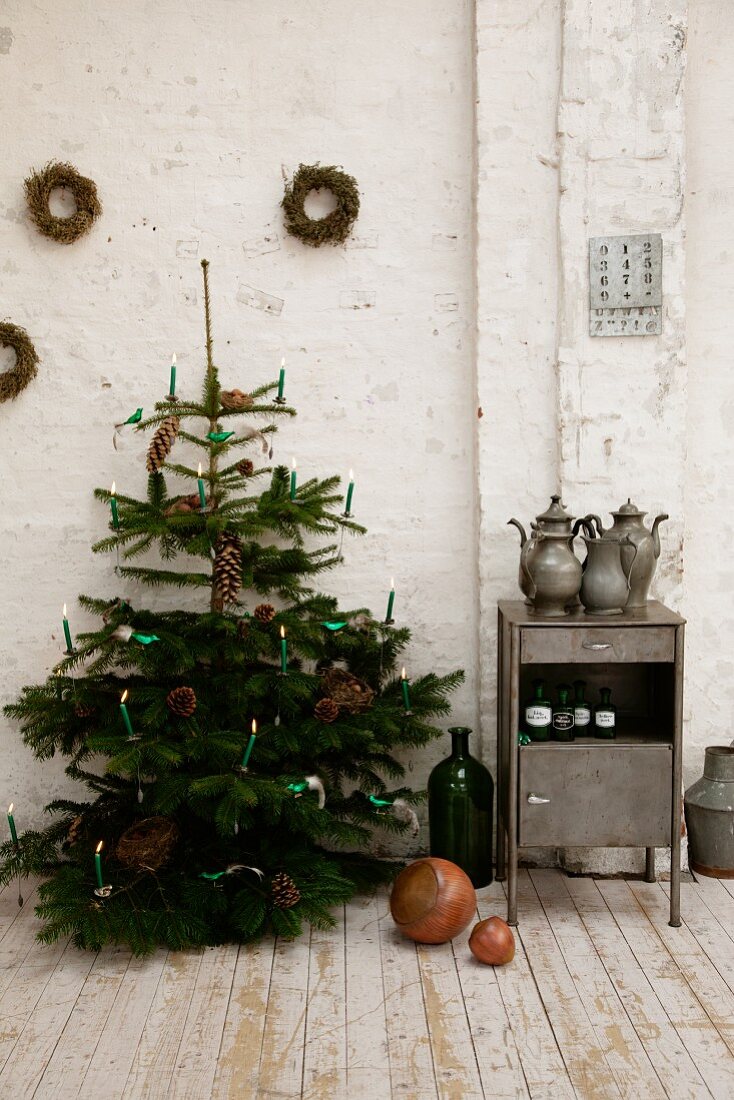 Christmas tree with lit candles next to small vintage cabinet against white wall in rustic atmosphere