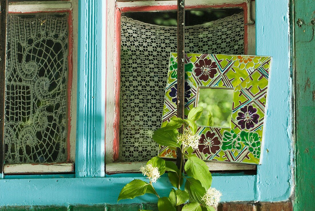 Small mirror with broad floral frame and brightly painted window frame with crochet curtains