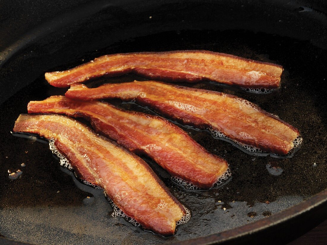 Vier Scheiben Frühstücksspeck werden in Pfanne gebraten