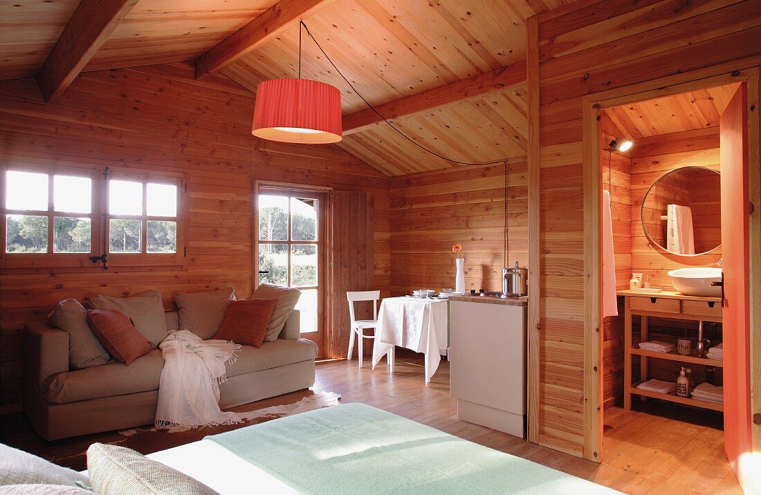 Offener Wohnraum in gemütlichem Holzhaus; eine zentrale Hängelampe sorgt für warmes Licht