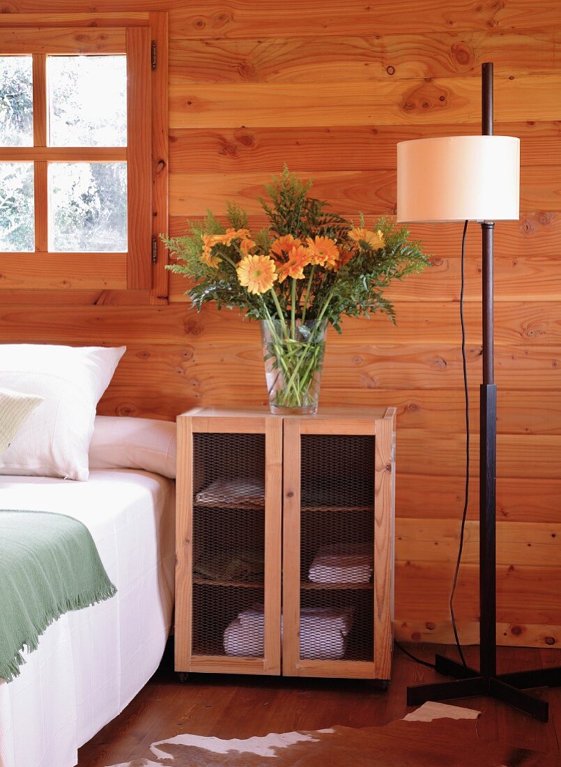 Glorious bouquet of orange flowers on small cupboard with wire mesh doors as bedside table next to double bed