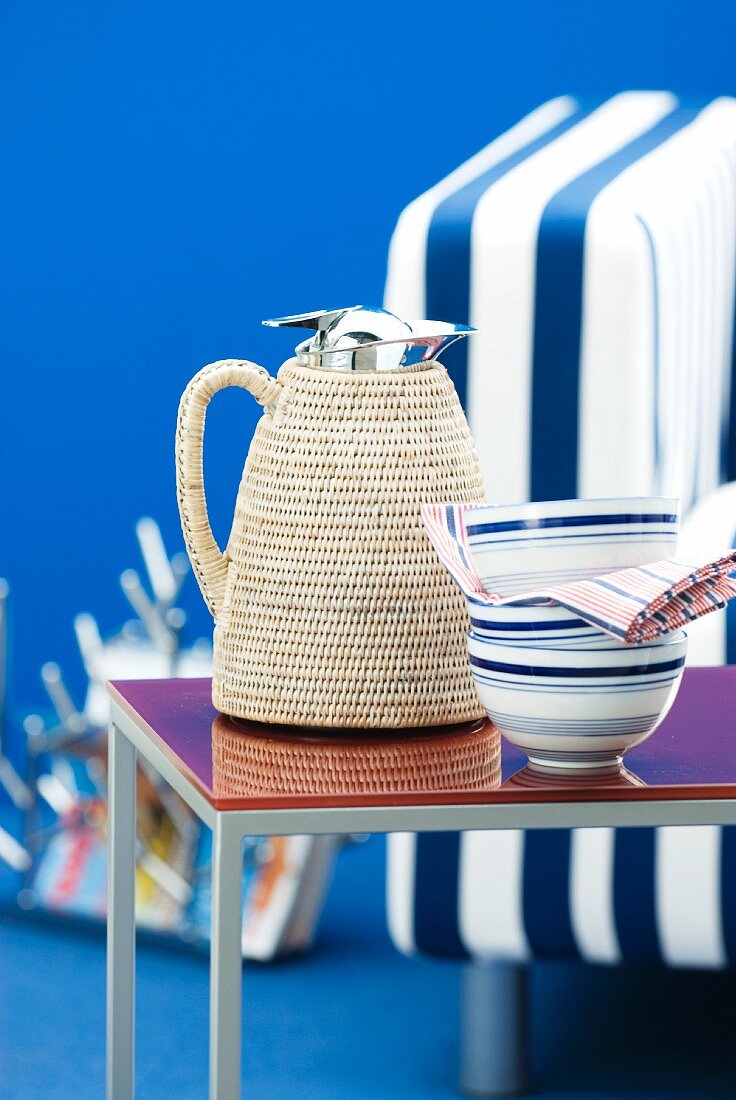 Thermos jug with raffia cover next to china bowls on side table