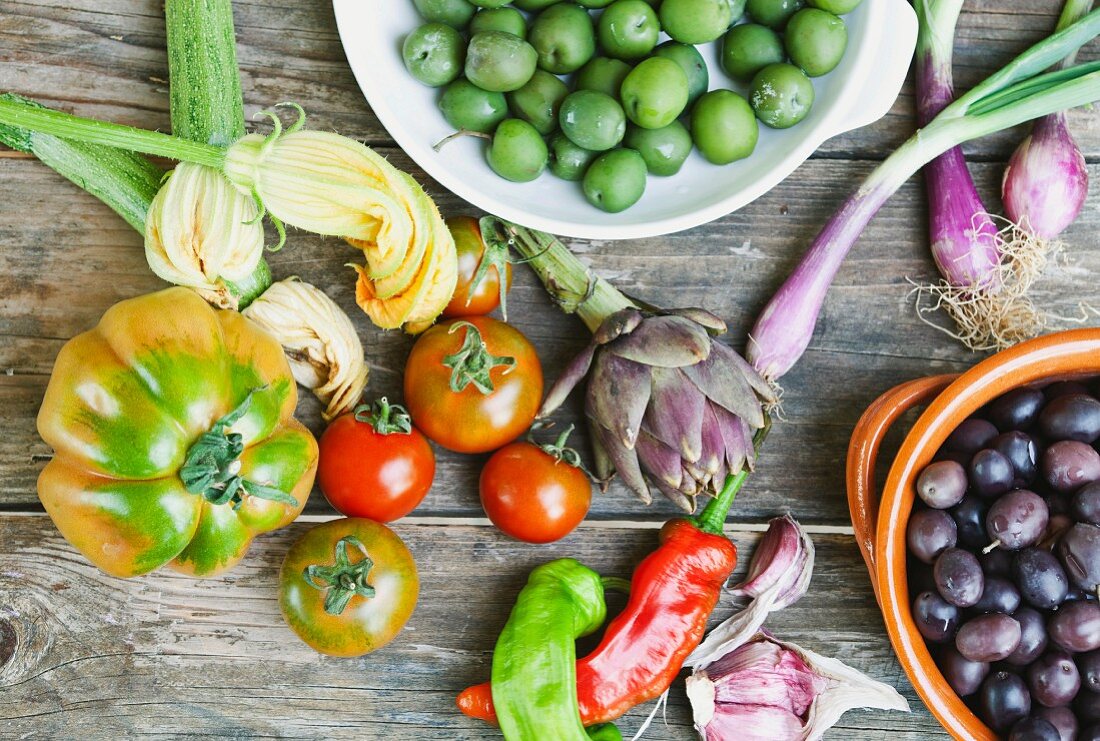 Two bowls of olives, spring onions, tomatoes, garlic, pepper, courgette flowers and artichokes on a wooden table