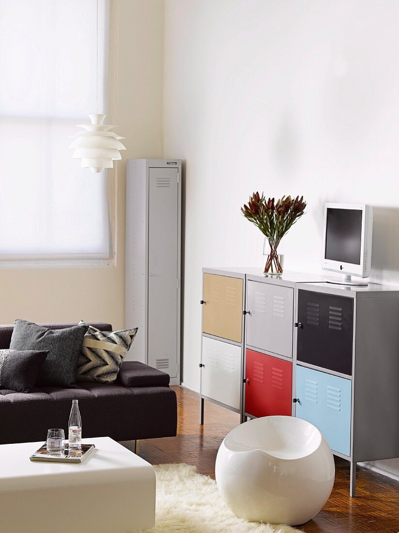 White, spherical plastic stool in front of half-height, locker-style, multicoloured metal cabinet and black designer sofa in corner of living room