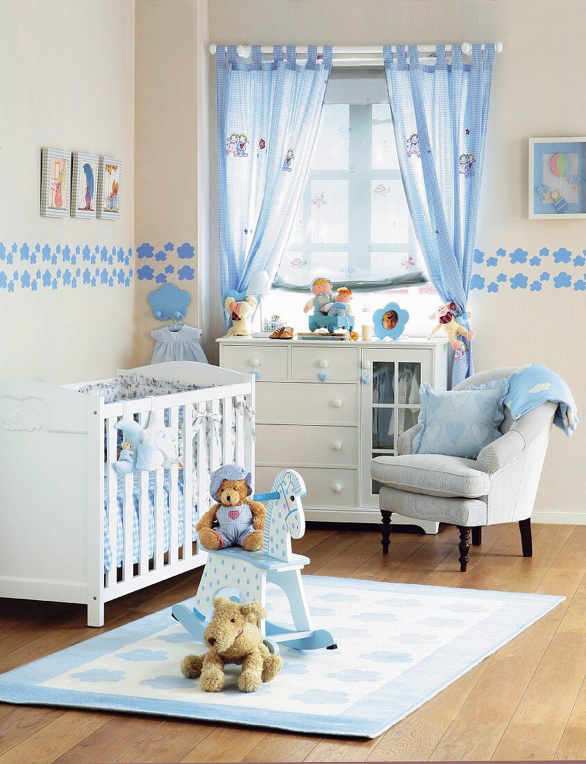 Play mat in front of white cot and armchair next to half-height chest of drawers below window in country-style nursery