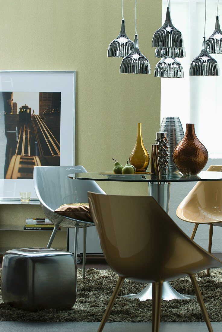 Metallisch farbene Schalenstühle an rundem Tisch mit Vasensammlung unter Hängeleuchtegruppe mit Metallschirm