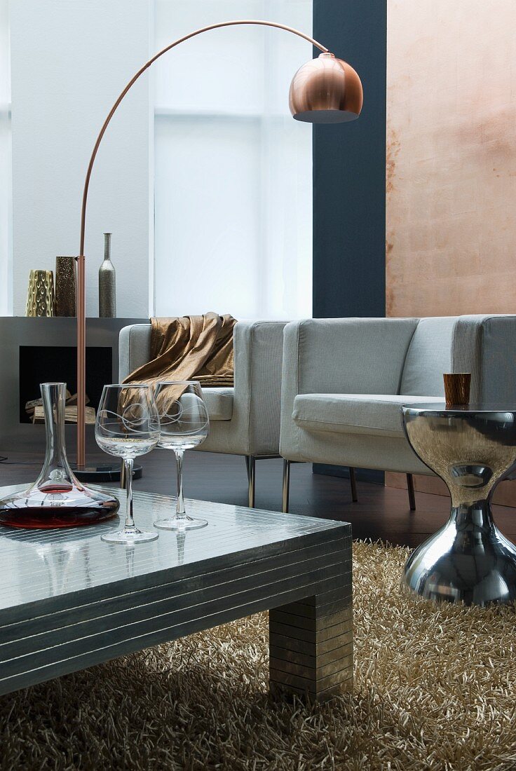 Wine decanter and glasses on low, metal coffee table in front of modern armchairs and copper-coloured arc lamp