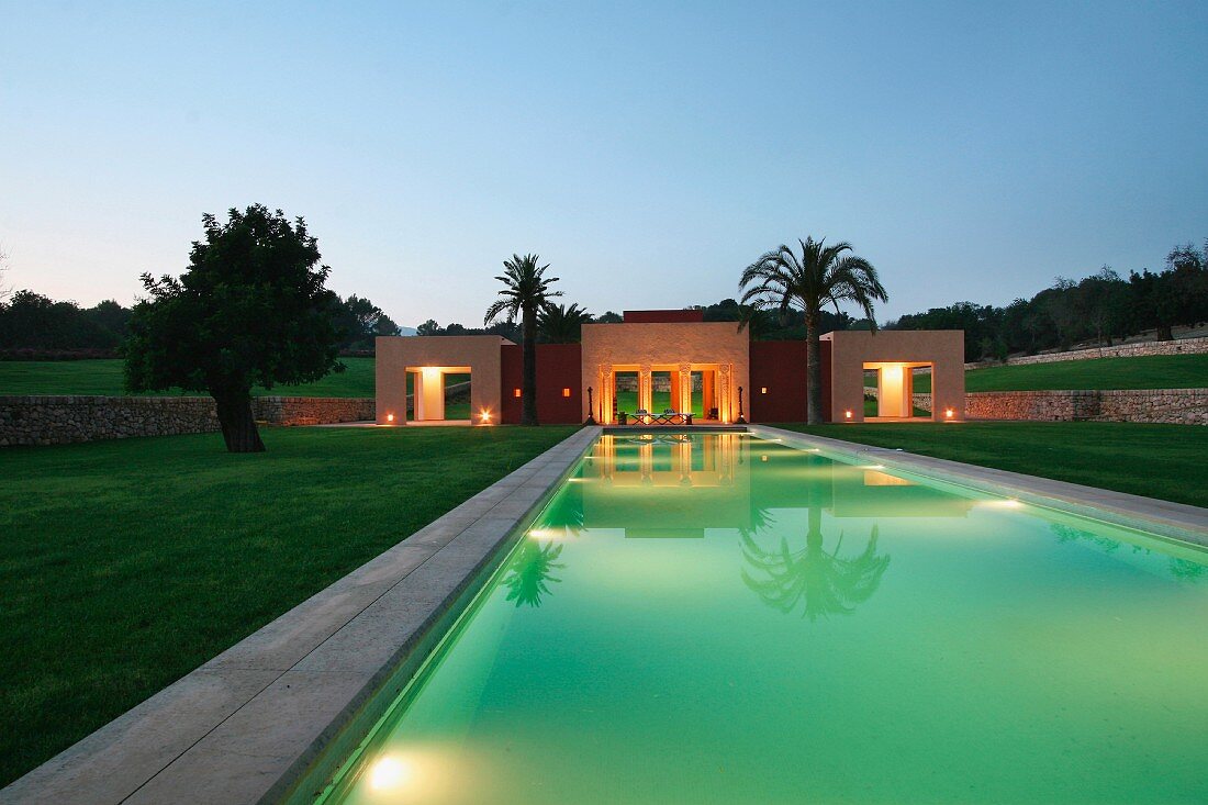 Ein langer, beleuchteter Pool mit mediterranem Poolhaus bei Nacht