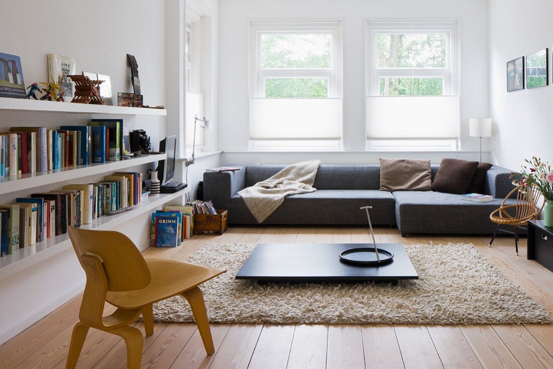 Low, wooden Bauhaus chair in front of floor-level, dark coffee table on flokati-style rug and grey designer sofa below window