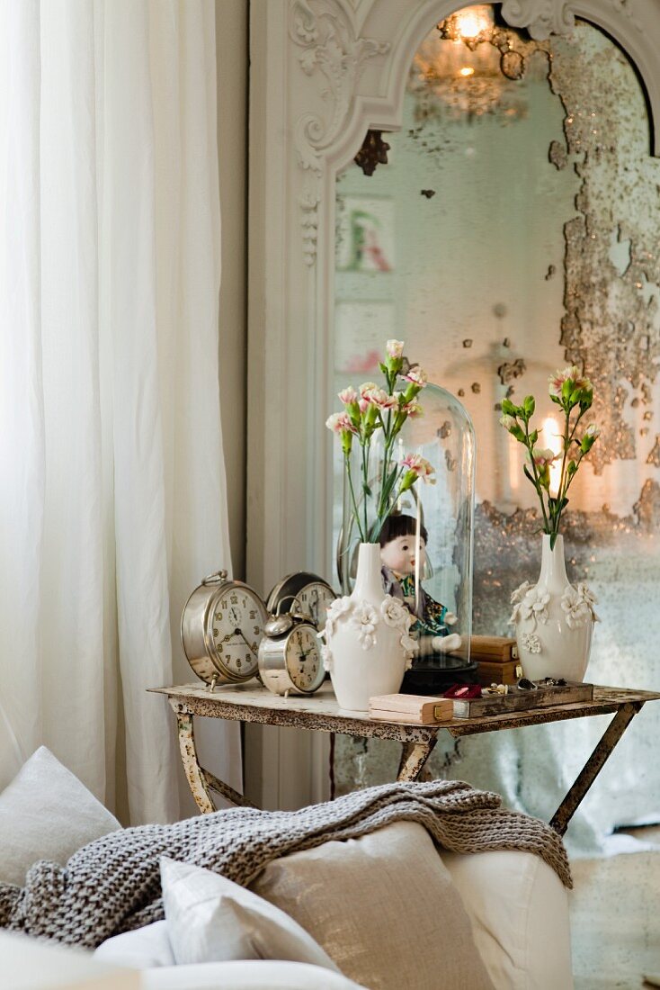 Alarm clocks, doll and vase of flowers on vintage side table next to mirror on wall