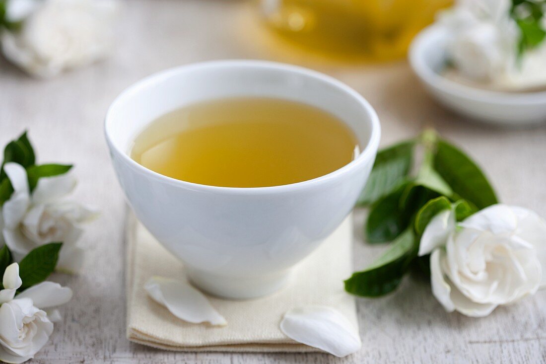 Eine Teeschale mit grünem Tee umgeben von Gardenienblüten