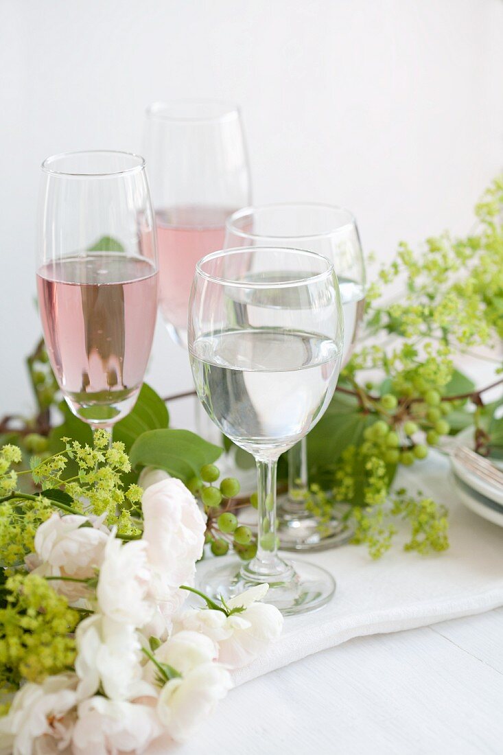Prosecco mit Rosensirup und Wasser auf gedecktem Tisch mit Rosen, Schleierkraut und Ranken