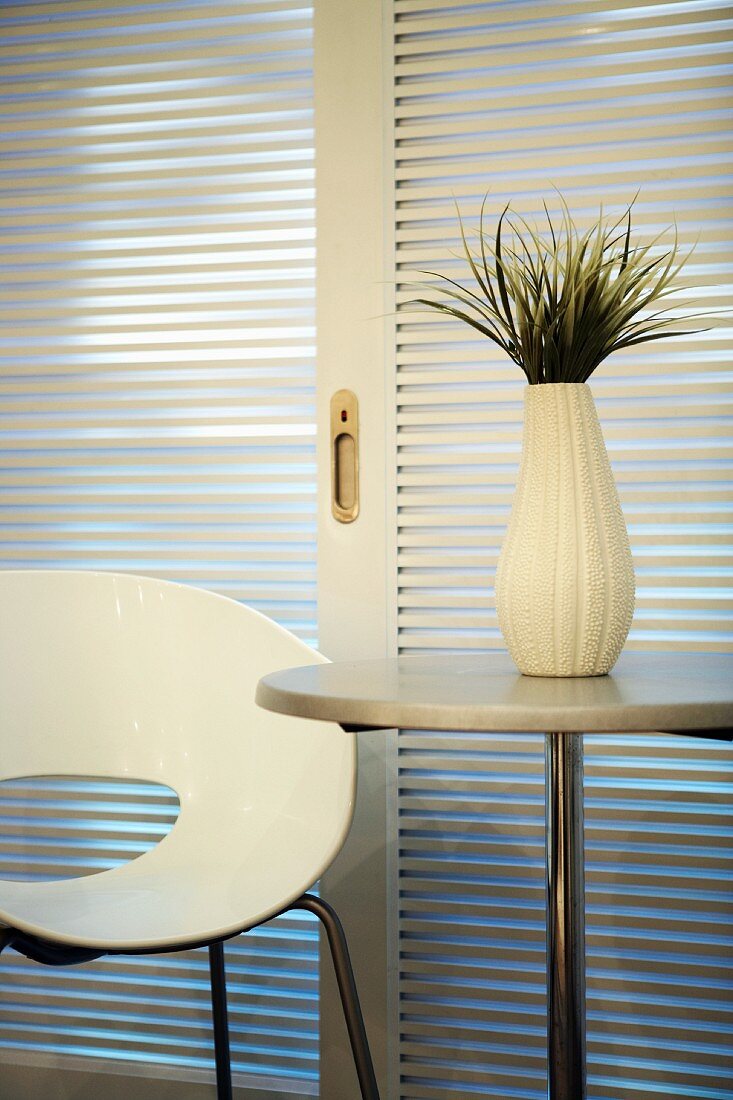 White designer chair next to white vase on round table in front of sliding blinds