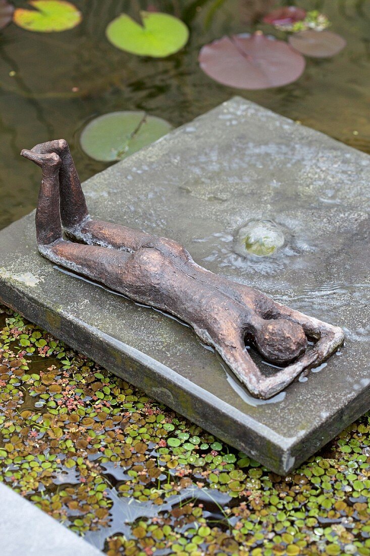 Reclining bronze figure on bronze raft in pool amongst duck weed and lily pads