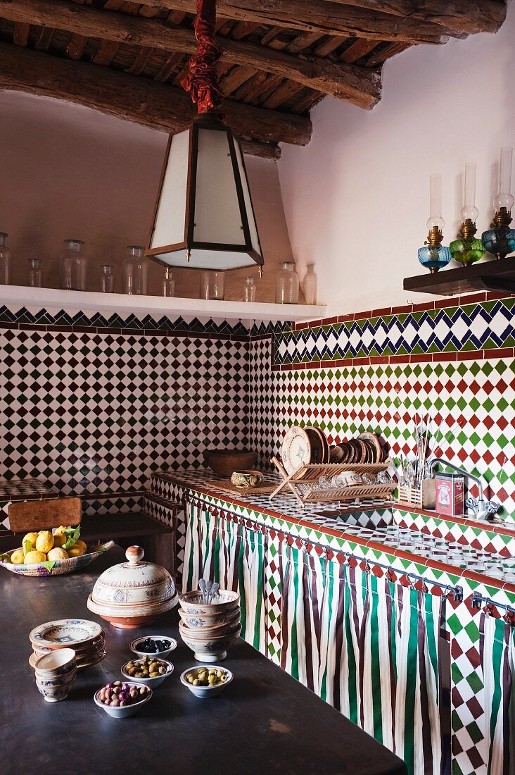 Corner of Oriental kitchen with mosaic-tiled sink unit and walls; shelves of oil lamps and glass vessels form border between tiles and plain painted walls; full bowls on wooden table in foreground