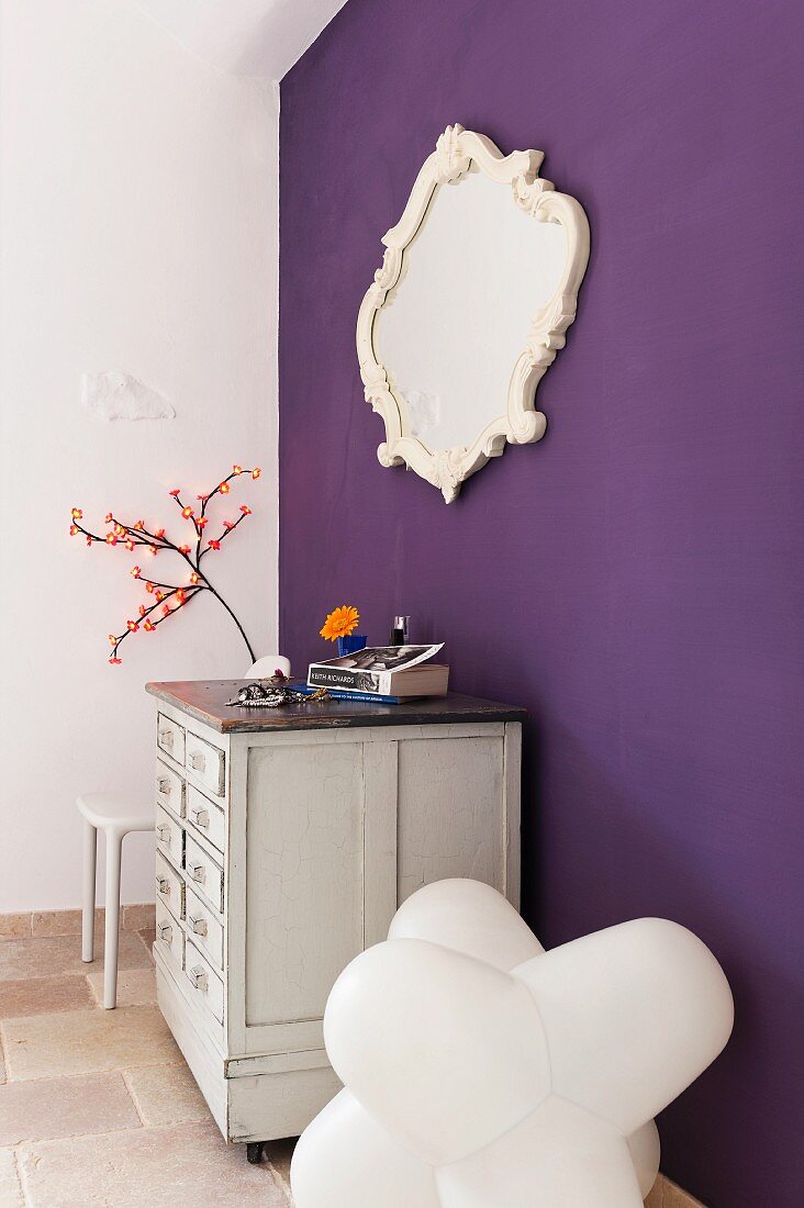 White commode, framed mirror and designer chair by a violet wall