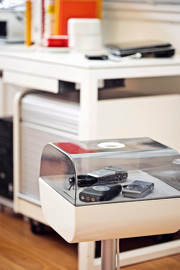 Various cameras in rounded, white plastic display case with plexiglass cover; blurred workstation in background