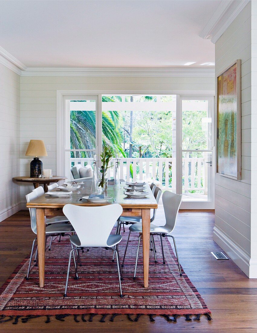 Airy dining area with large wooden table and white chairs; in the background a sunny balcony with a view of a large palm tree