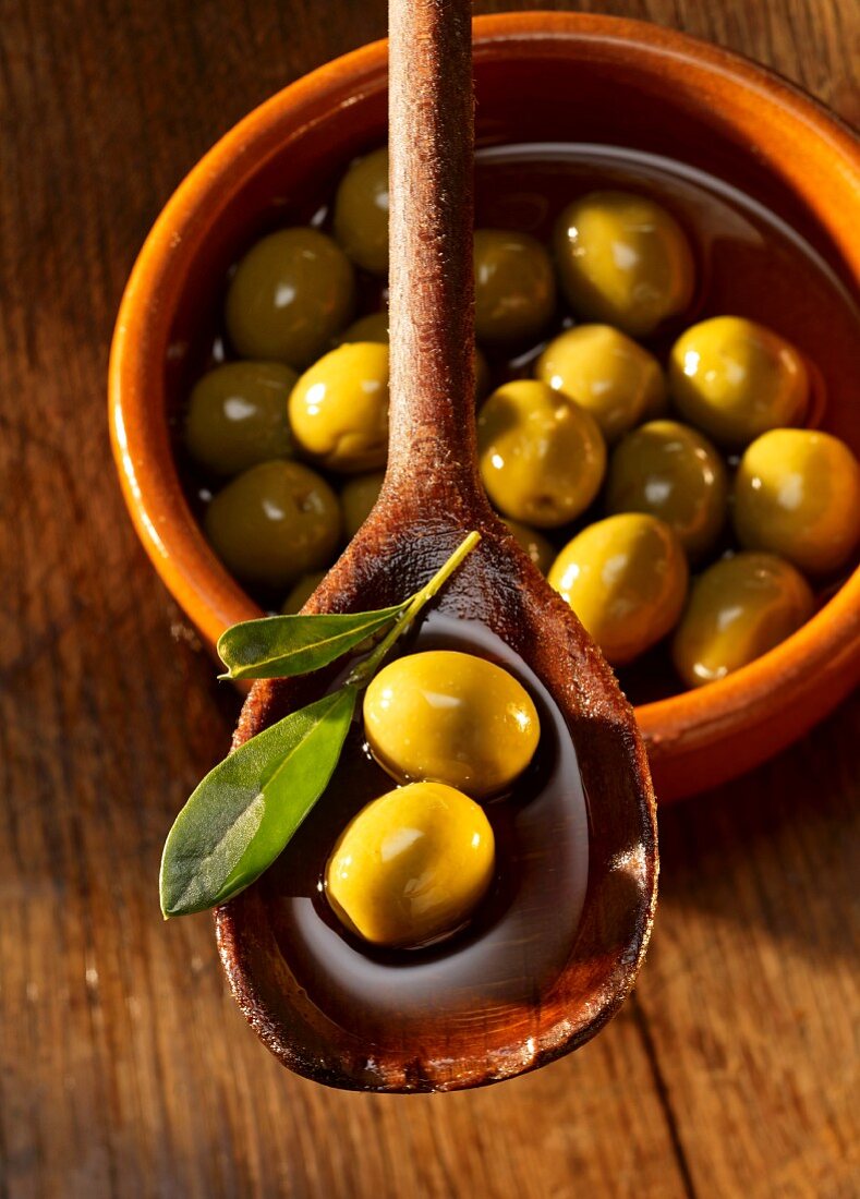 Grüne Oliven in Olivenöl auf Holzlöffel und in Tonschale