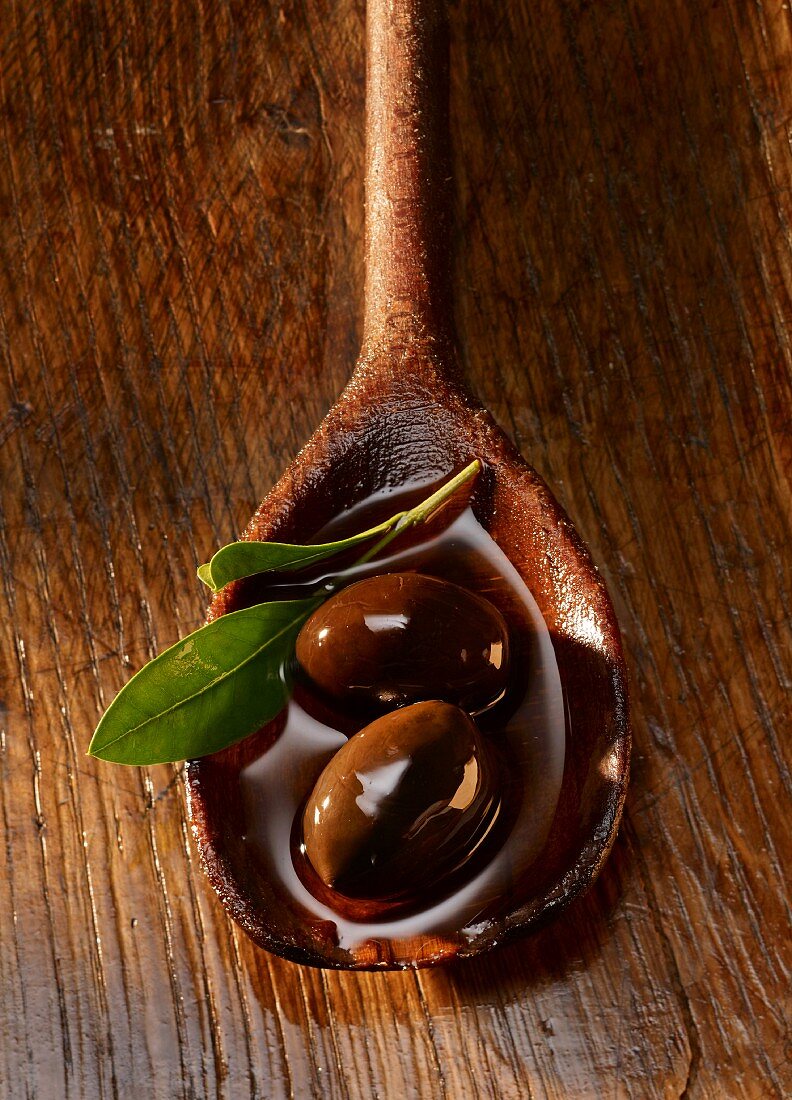 Black olives and an olive leaf in olive oil on a wooden spoon