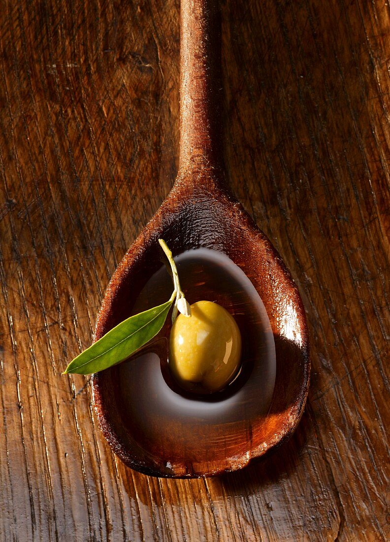 A green olive and an olive leaf in olive oil on a wooden spoon
