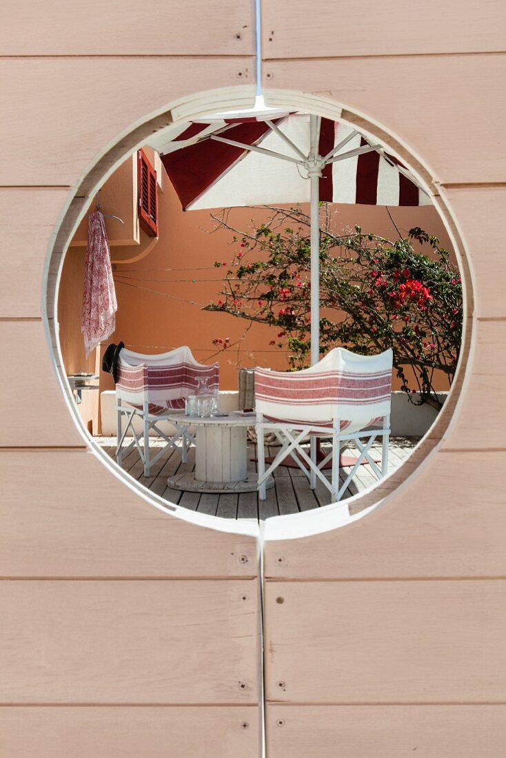 Blick durch Bullauge einer Holztür auf Holzterrasse mit bequemen Regiestühlen und Sonnenschirm