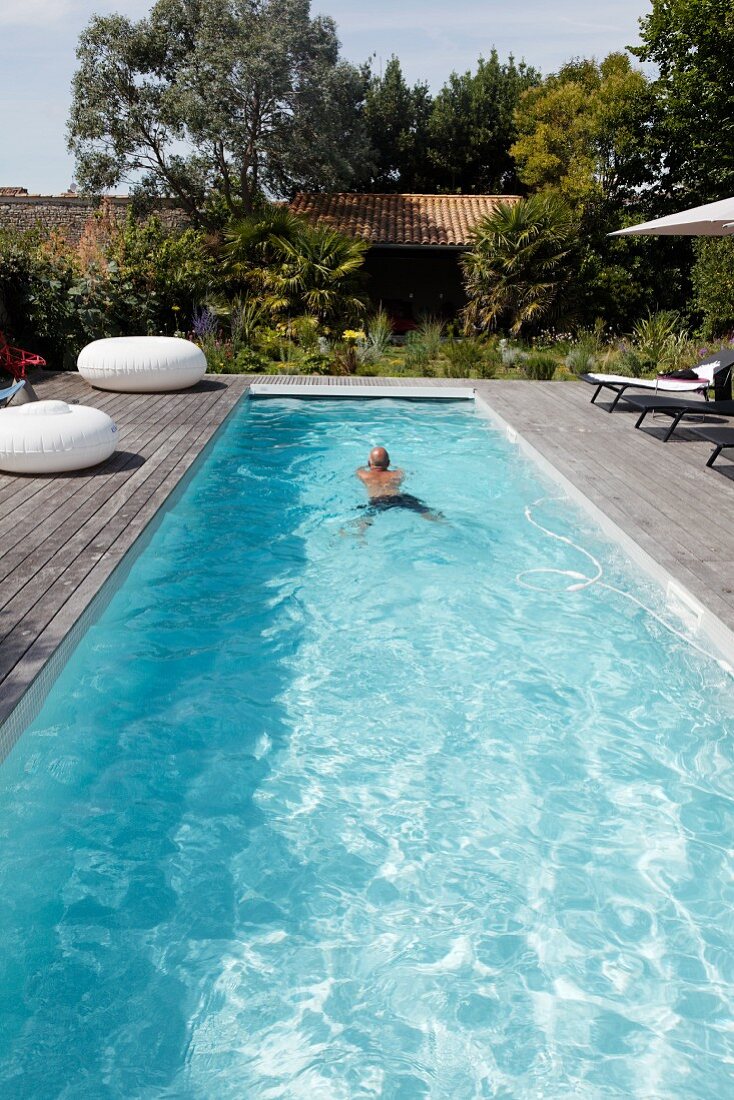 In länglichem Pool schwimmender Mann auf sonniger Holzterrasse mit verschiedenen Sitzgelegenheiten